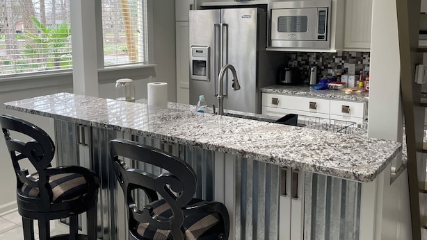 Una cocina moderna con armarios blancos, encimera de granito, electrodomésticos de acero inoxidable y dos taburetes negros.