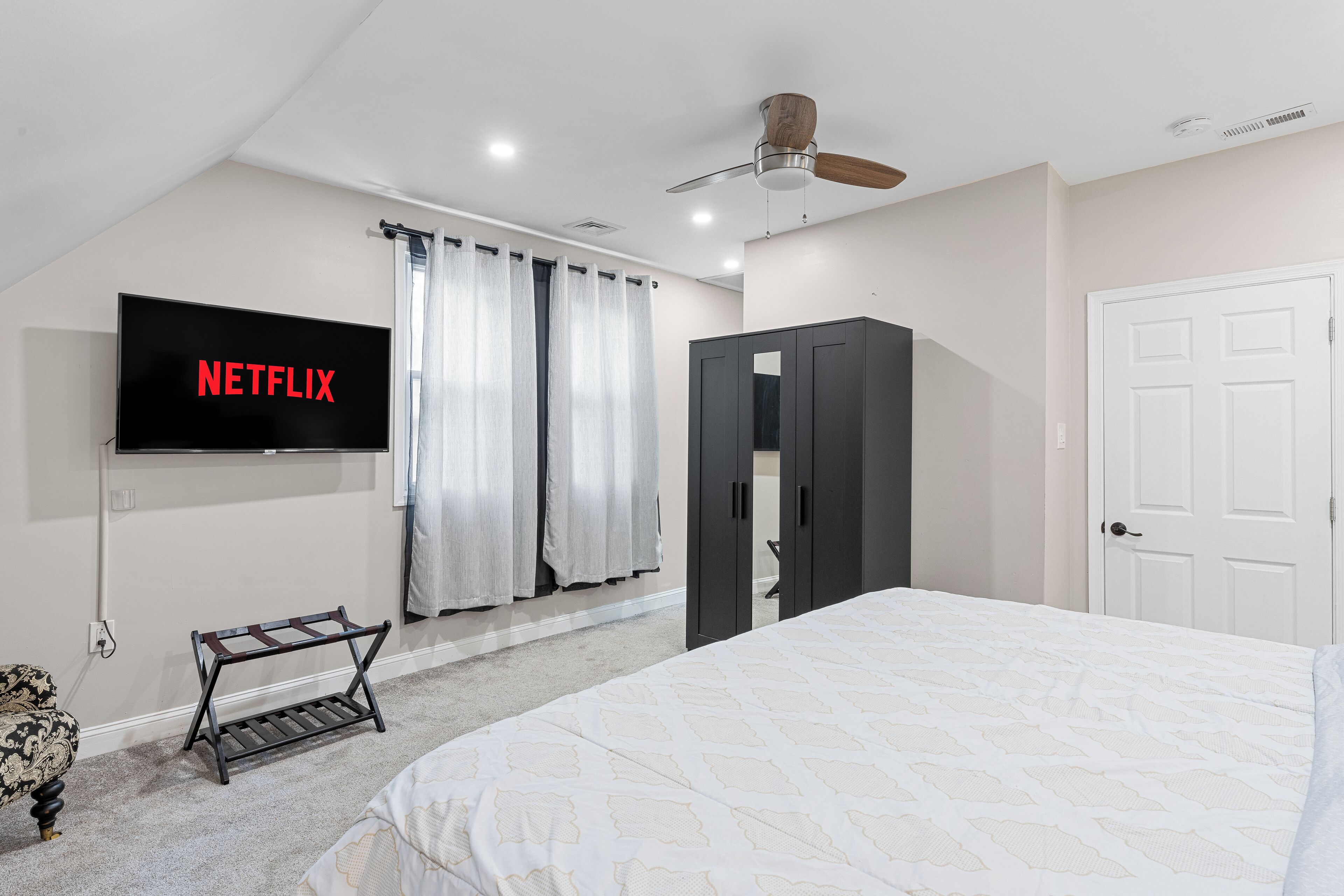 A bedroom with a bed, a TV mounted on the wall, a wardrobe, and a ceiling fan.