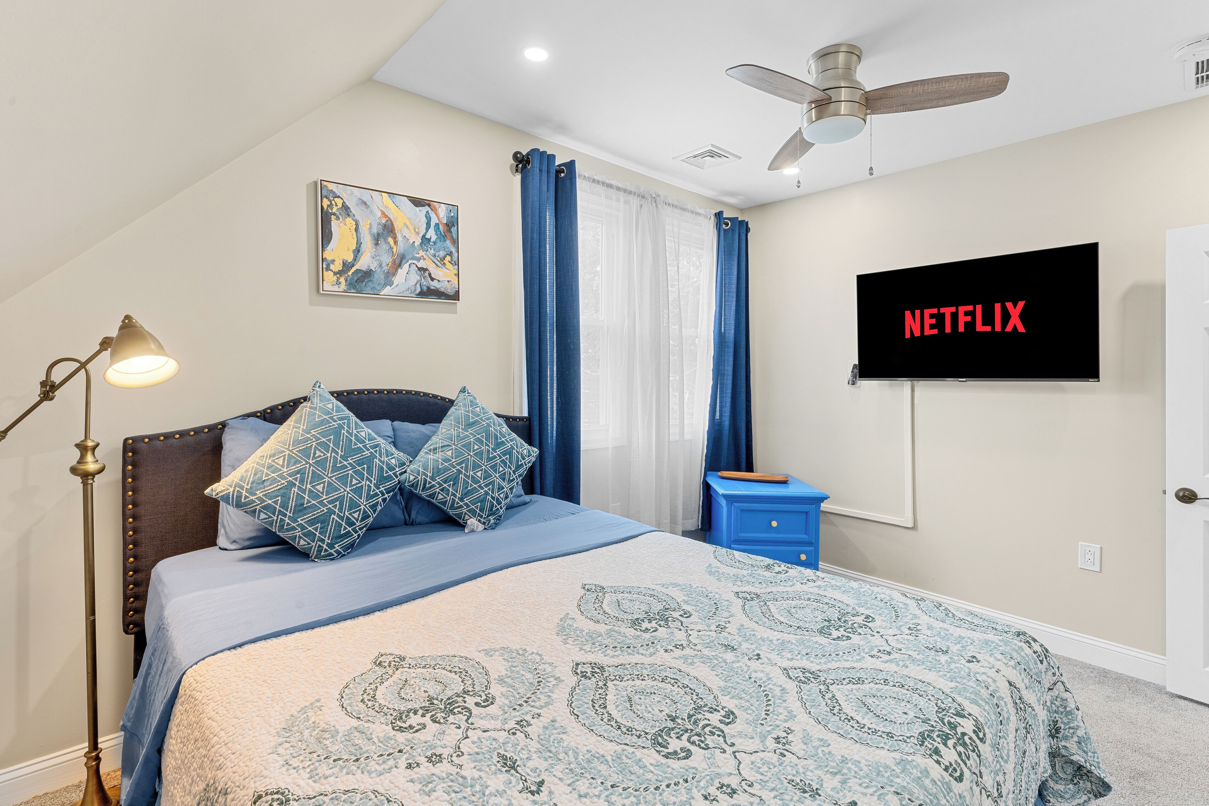 A bedroom with a bed, a ceiling fan, a TV displaying 'NETFLIX', and a painting on the wall.