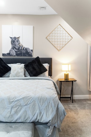 A bedroom with a bed, a nightstand, a lamp, a tiger painting, and a geometric wall decoration. - Tudor Lux (East Lansdowne)