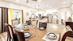 A dining area with a wooden table set for six, surrounded by chairs. A ceiling fan is above, and the room has a modern kitchen in the background. - Tudor Lux (East Lansdowne)