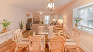 Un coin repas avec des chaises en osier et une table en verre, un lustre et une vue sur l’extérieur par une fenêtre munie de stores.