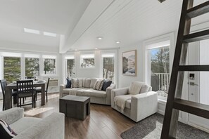 En rummelig opholdsstue med en stor sofa, et spisebord og flere vinduer med udsigt til omgivelserne.