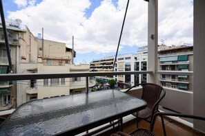 Un balcón con una mesa de vidrio y sillas de mimbre, con vista a un paisaje urbano con diversos edificios.