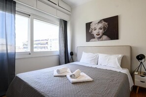Habitación de hotel con cama, ventana con cortinas, un cuadro colgado en la pared y una mesita de noche con lámpara.