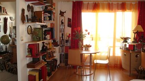 Ein gemütliches Zimmer mit Holzboden, einem Esstisch mit Stühlen, einem Bücherregal voller Gegenstände und einem Fenster mit roten Vorhängen.