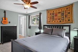 Ein Schlafzimmer mit einem Bett, einem Deckenventilator, einer Gitarre, einem Schminktisch, einem Nachttisch, einer Lampe und einem Fenster.