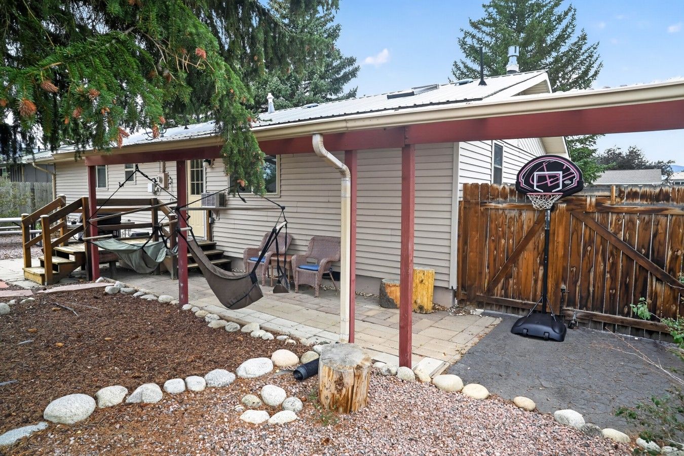 A residential backyard with a swing set, a basketball hoop, and a covered patio area.