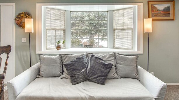 Ein Zimmer mit einem großen Fenster, einem Sofa mit Kissen, einem kleinen Tisch mit Pflanze und Wandlampen.