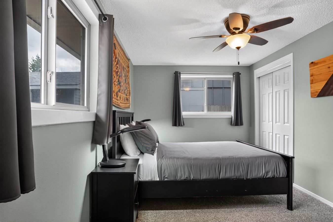 Une chambre à coucher avec un lit, une table de chevet, une lampe, une fenêtre et un ventilateur de plafond.
