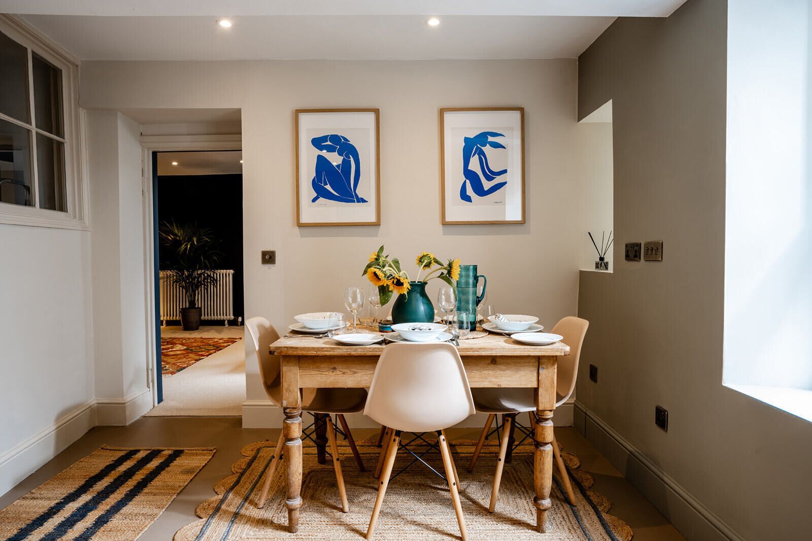 A dining area with a wooden table set for four, two framed abstract paintings above, and a view of a garden through an open door.