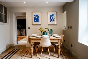 Un coin repas avec une table en bois dressée pour quatre personnes, deux tableaux abstraux encadrés au-dessus, et une vue sur un jardin par une porte ouverte.