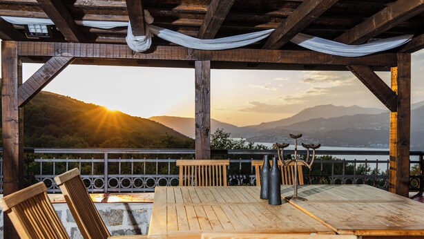 A wooden outdoor seating area with a table and chairs, overlooking a scenic view of mountains and a body of water during sunset. - Authentic Mediterrannean Villa Ancient Mill with Swimming Pool and Sea View (Luštica)
