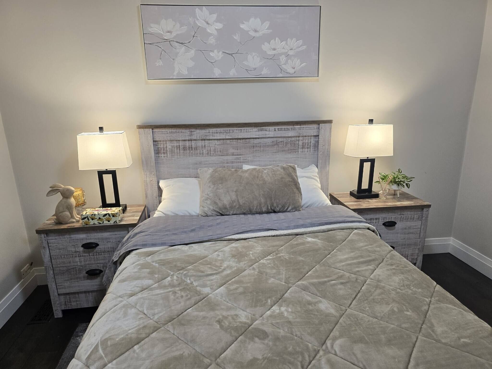 Un dormitorio con una cama, dos mesitas de noche, una lámpara, un conejo decorativo, un cuadro enmarco y un conjunto de cajones.