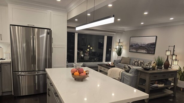 A modern kitchen with a stainless steel refrigerator, a marble island with fruit, and a dining area with a gray sofa and table. - Comfortable City Home (Mississauga)