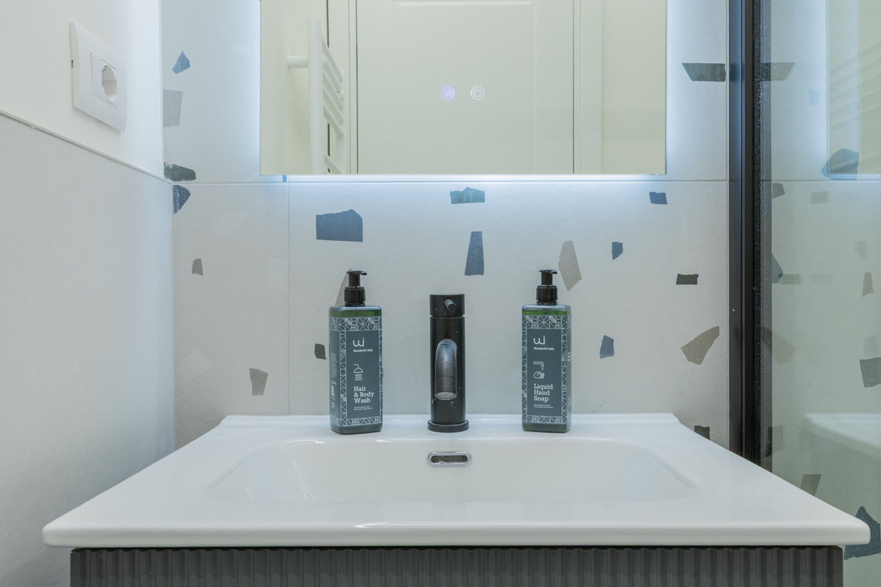 Un baño con un lavamanos blanco, dos botellas de jabón para cabello y cuerpo, y un espejo encima.