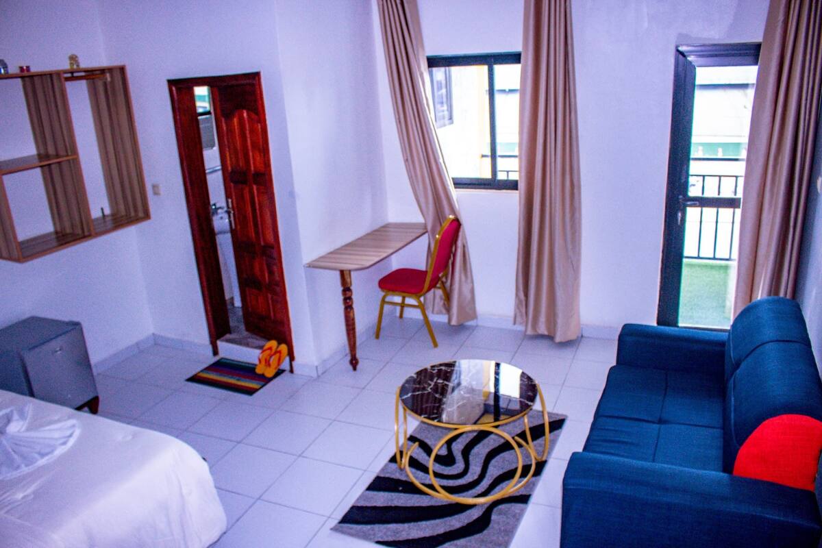 A hotel room with a bed, a blue sofa, a small table, a chair, and a window with curtains.