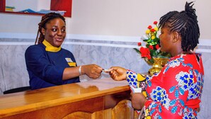 A reception desk with a wooden counter, a vase with flowers, and a person wearing a blue shirt. - WEST HOTEL DOUALA  (Bonaberi)