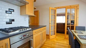 A compact kitchen with a gas stove, oven, and wooden cabinets. A dining area with a table and chairs is visible through an open door. - Lochiel Apartment Self Catering (Fort William)
