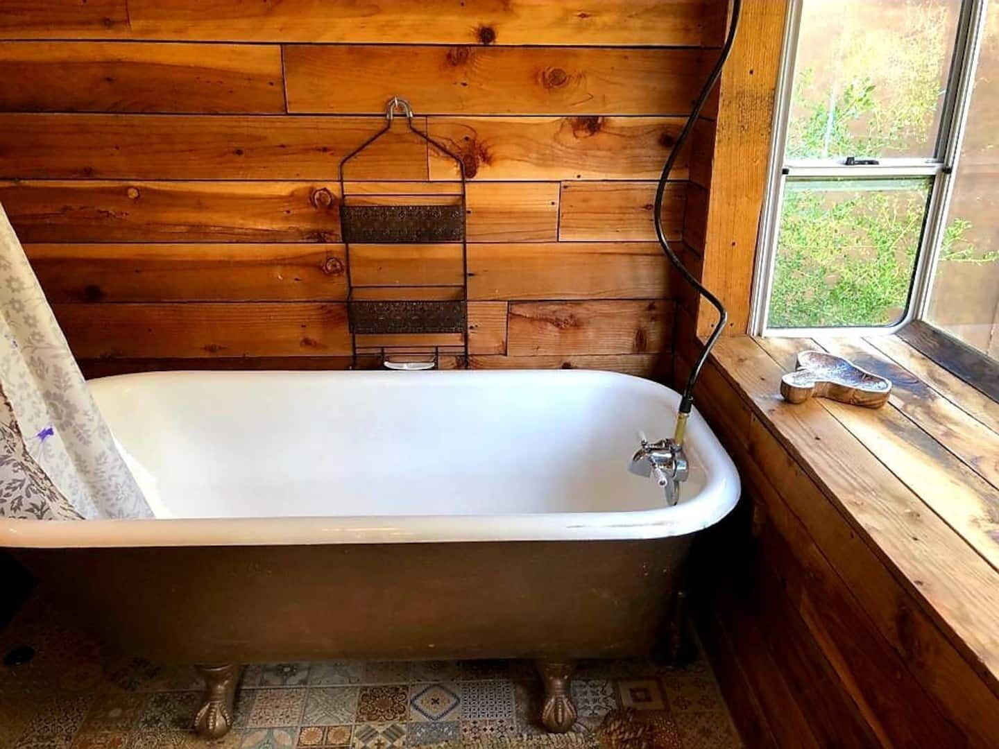 A wooden cabin bathroom with a white bathtub, a window with a view of greenery, and a towel rack.