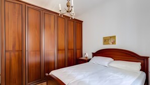 Quarto com guarda-roupa de madeira, cama com roupa de cama branca, mesa de cabeceira, abajur e um ornamento na parede.