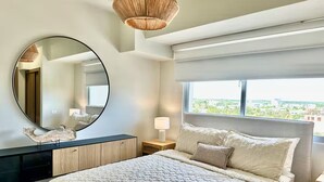 Une chambre avec un grand lit, un miroir rond, une table de chevet avec une lampe, et une vue sur la verdure à l’extérieur.