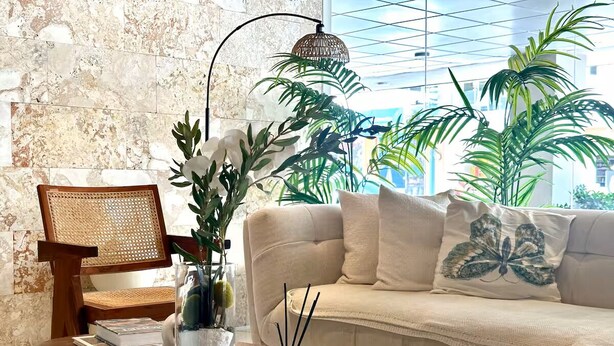 Un salon moderne avec une table basse en bois, un fauteuil en rotin, un canapé beige agrémenté de coussins décoratifs et une grande fenêtre donnant sur des immeubles.