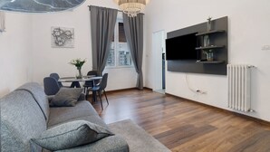 A modern living room with a grey sofa, dining table, chairs, a chandelier, a flat-screen TV, and a radiator. - Apartment 'Home Experience - Perla Grigia' with Air Conditioning (Bari)
