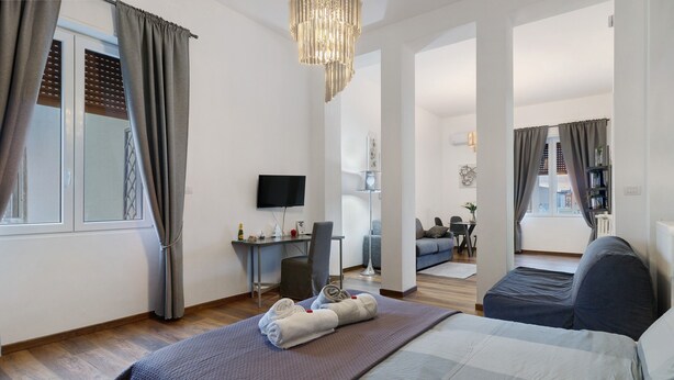 A modern hotel room with a large bed, a desk, a TV, and a view of the city. - Apartment 'Home Experience - Perla Grigia' with Air Conditioning (Bari)