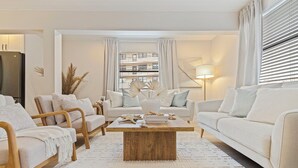 Una sala de estar moderna con un sofá beige, dos sillas de madera, una mesa de centro y un ventanal con persianas.
