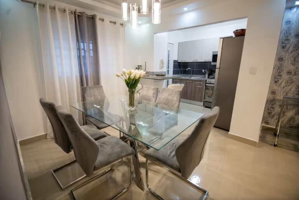 Uma sala de jantar moderna com mesa de vidro e cadeiras estofadas cinzas, um vaso com flores brancas, e uma cozinha ao fundo.