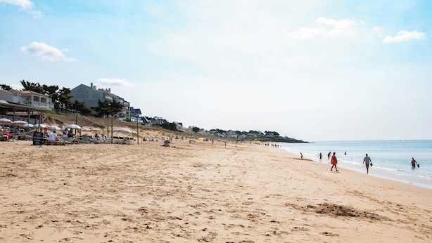 A wide sandy beach with footprints, a few people walking, and buildings in the background.