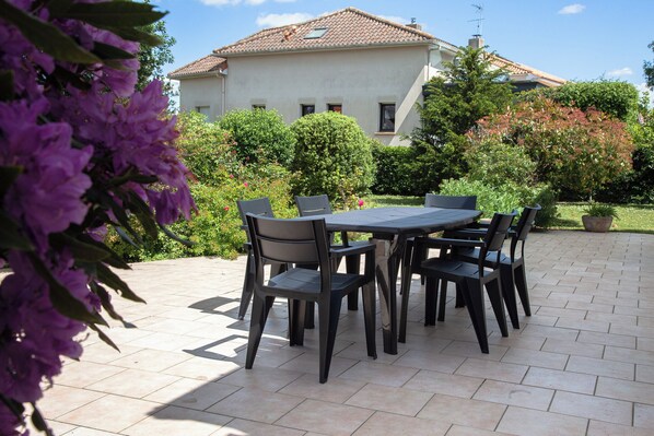 An outdoor patio with a table and chairs, surrounded by greenery and a building in the background. - Family house 8/12pers in Pornichet - Garden 700 m² - Walking distance to beach (600 m) (Pornichet)