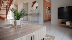 A modern living room with a wooden coffee table, a potted plant, a flat-screen TV, and a kitchen area in the background.