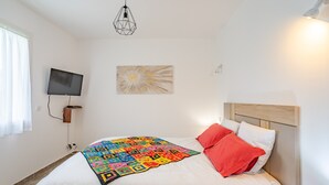 Ein ordentlich bezogenes Bett mit farbenfroher Steudecke, ein an der Wand montierter Fernseher und eine moderne Pendelleuchte.