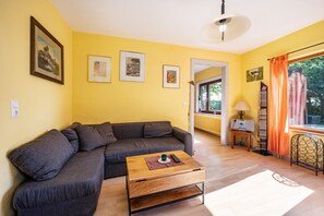 Un salon avec un canapé, une table basse et une lampe. Des tableaux encadrés ornent le mur, et une fenêtre est agrémentée de rideaux.