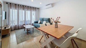 A modern living room with a wooden dining table, white chairs, a blue sofa, and a small side table with a vase of flowers. - LA SOLANA Roses / Roses - INTERimmo (Rosas / Roses)