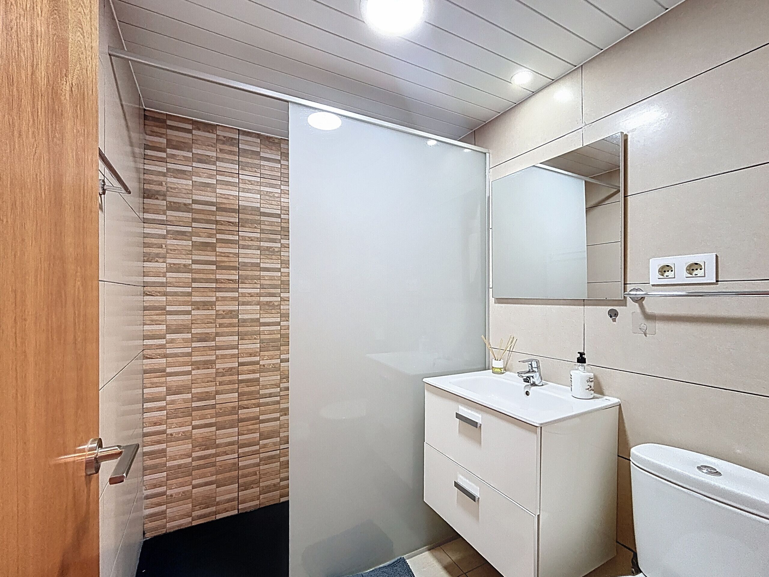 Een moderne badkamer met een houten deur, een douchecabine van glas, een witte wastafel met een spiegel erboven en een toilet.