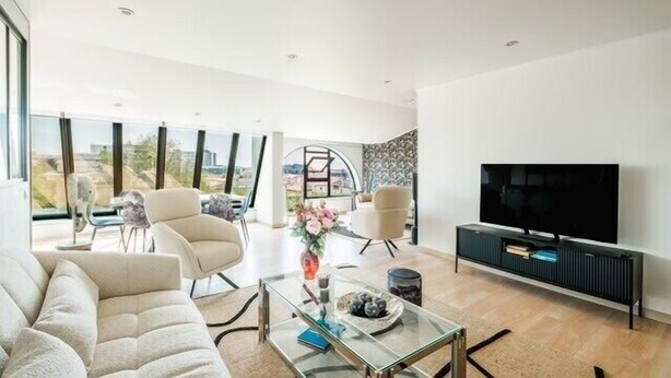 Un salon moderne avec un canapé, des fauteuils, une table basse en verre, un téléviseur et une vue sur la ville grâce à de grandes fenêtres.