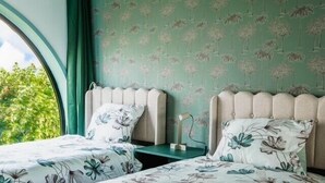 Une chambre avec deux lits, chacun recouvert d’une couvre-lit à motifs floraux et agrémenté d’oreillers assortis. Les murs sont recouverts d’un papier peint à motifs floraux verts, et une fenêtre offre une vue sur la verdure environnante.