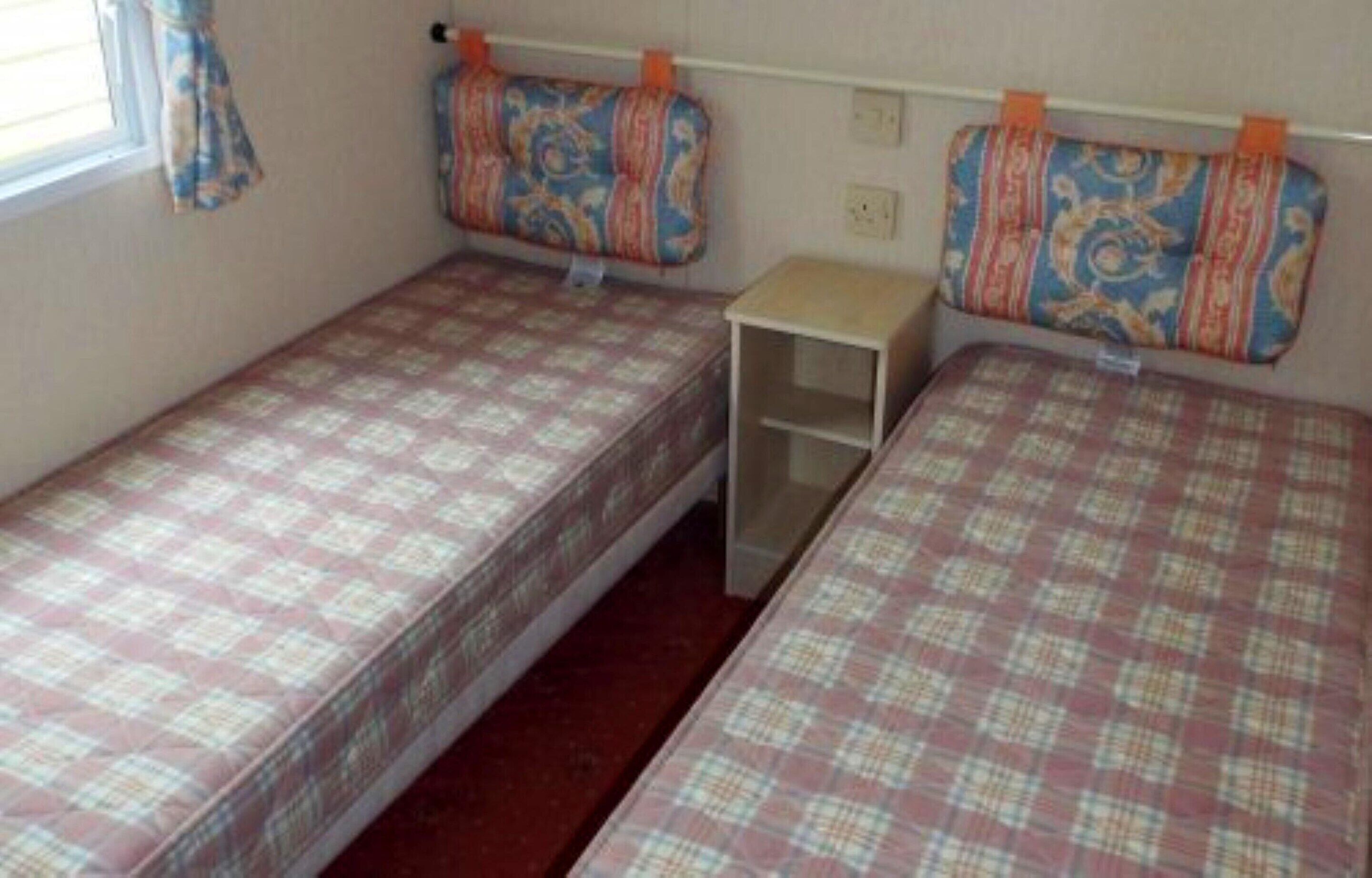 Una stanza modesta con due letti singoli, ognuno con una coperta a fantasia. Un tavolino è posizionato tra i letti, e una finestra con tende si trova sul lato sinistro.