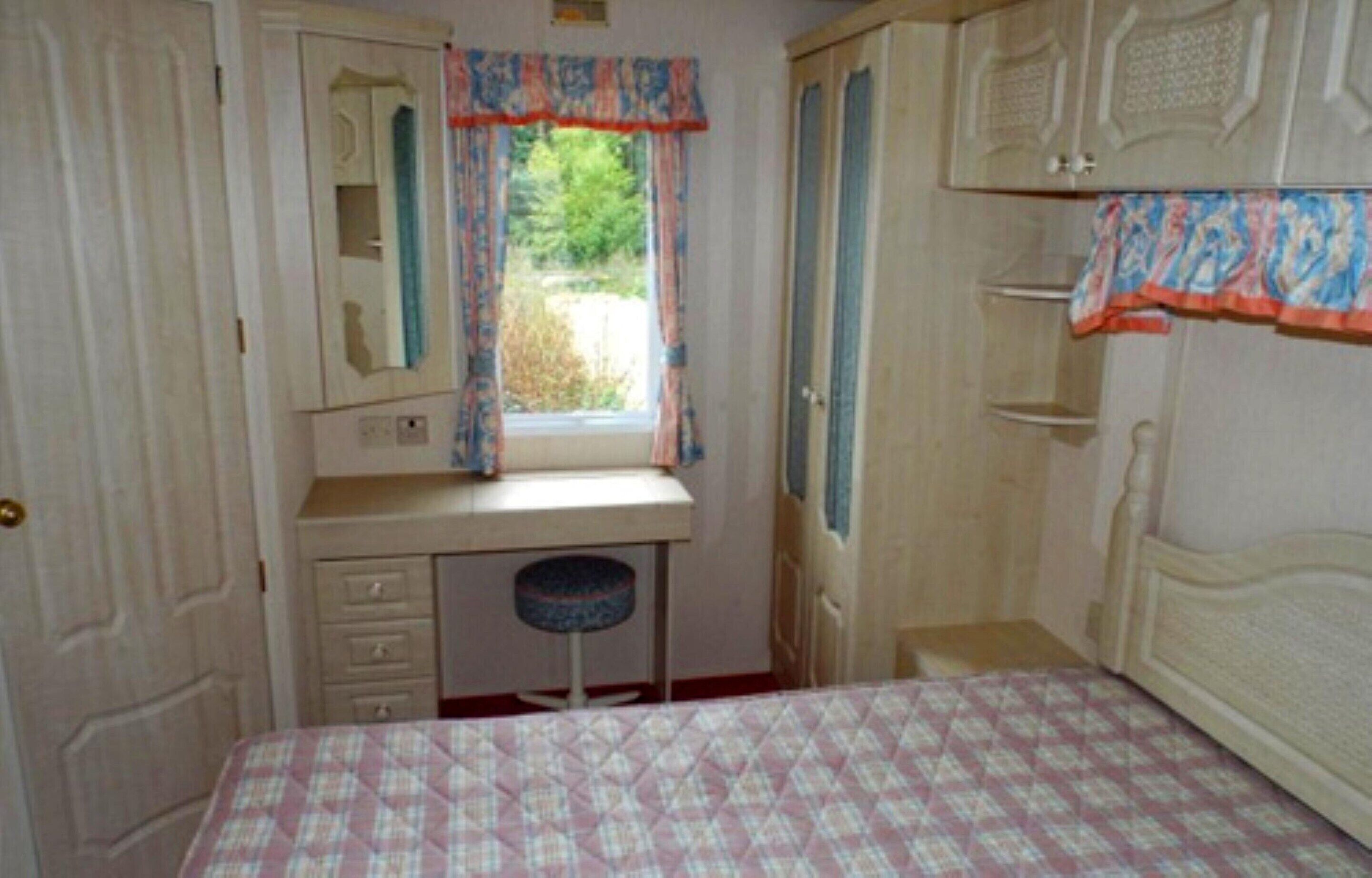 A bedroom with a bed, a desk, a chair, a window with curtains, and a wardrobe.