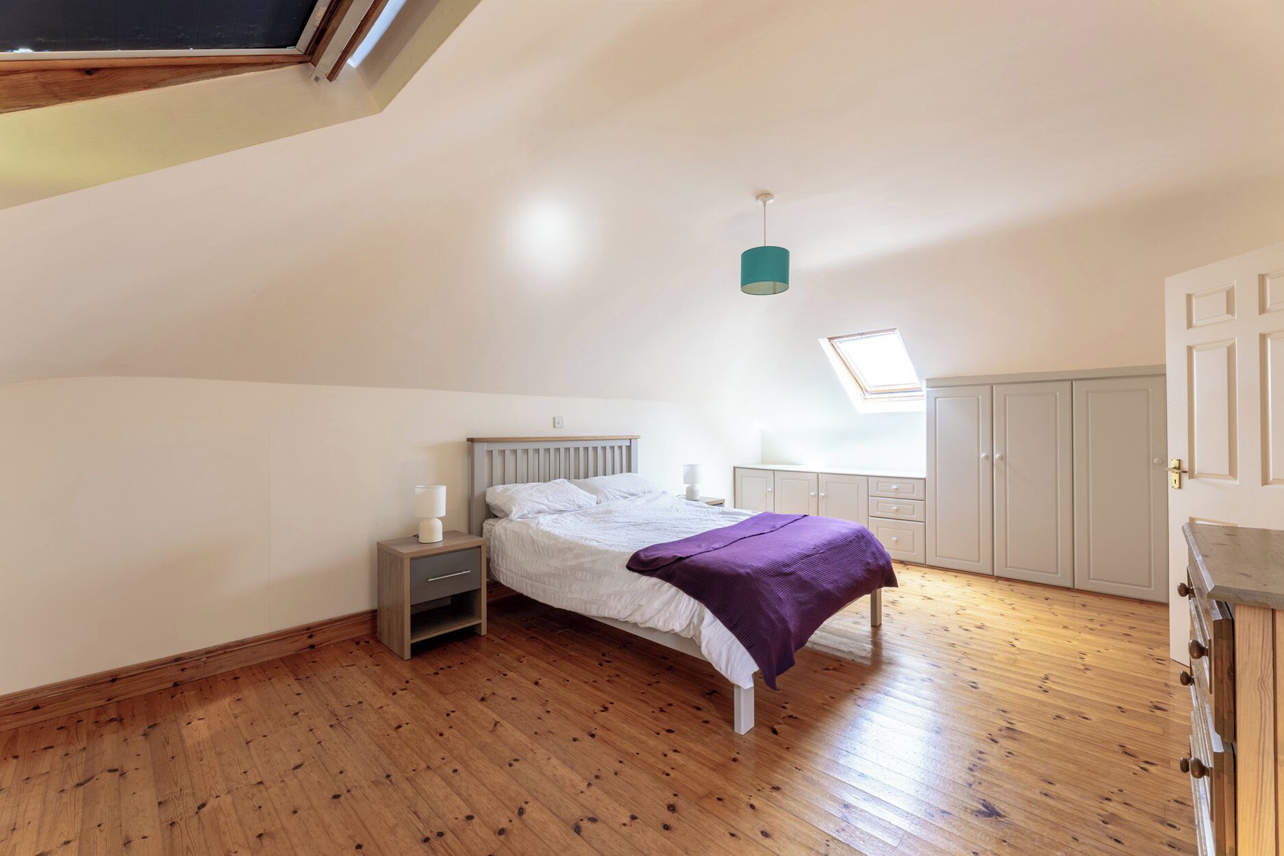 A bedroom with a sloped ceiling, a bed with a white headboard, a nightstand with a lamp, a wardrobe, and a skylight.
