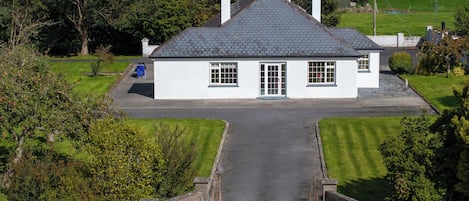 A white house with a grey roof, a front door, and a driveway leading to a gated entrance.