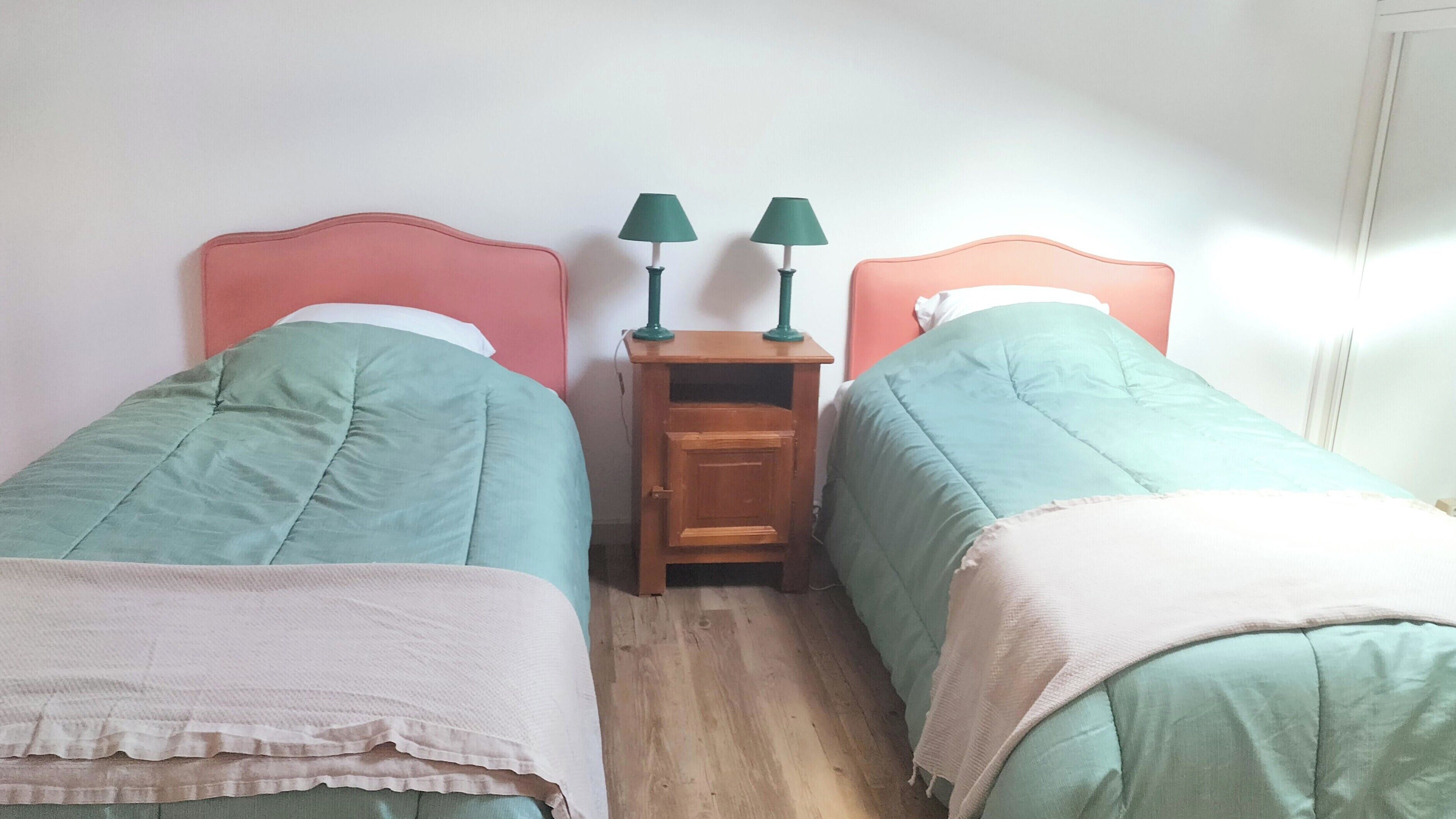 Un dormitorio con dos camas individuales, cada una con una colcha verde y un cojín blanco. Entre las camas hay una mesita de noche de madera con dos lámparas verdes.