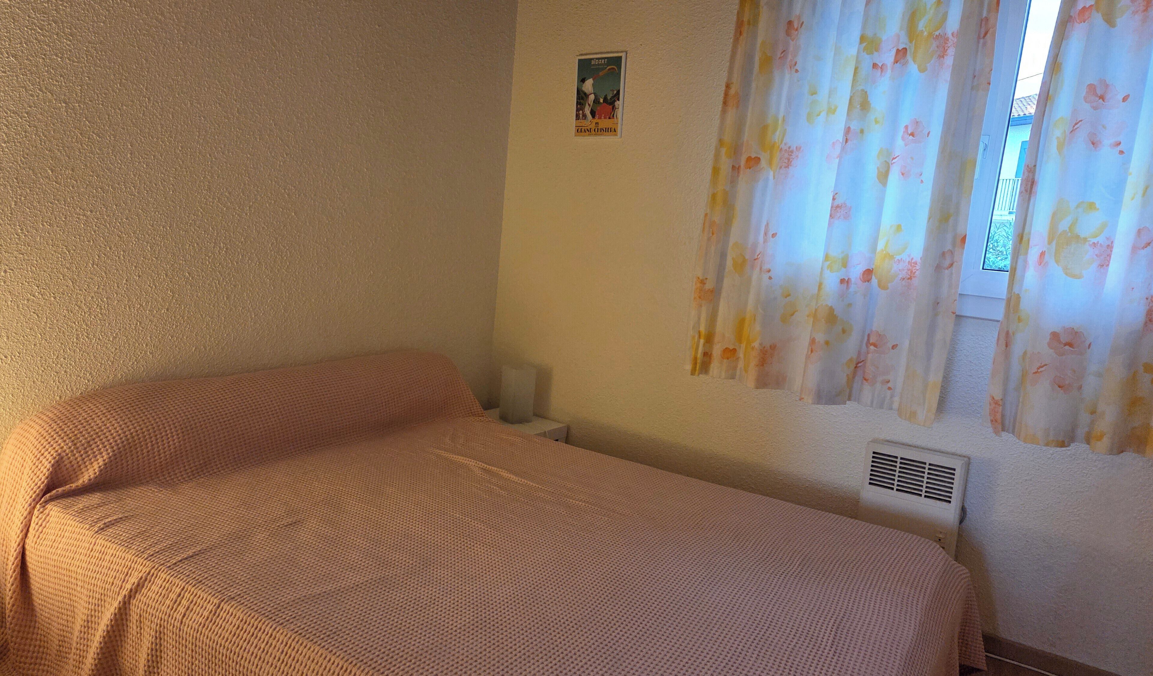 Una habitación con una cama, una mesita pequeña, un respiradero en la pared y un cartel.