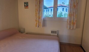 A bedroom with a bed, a window with curtains, a wall-mounted air vent, and a small poster on the wall.