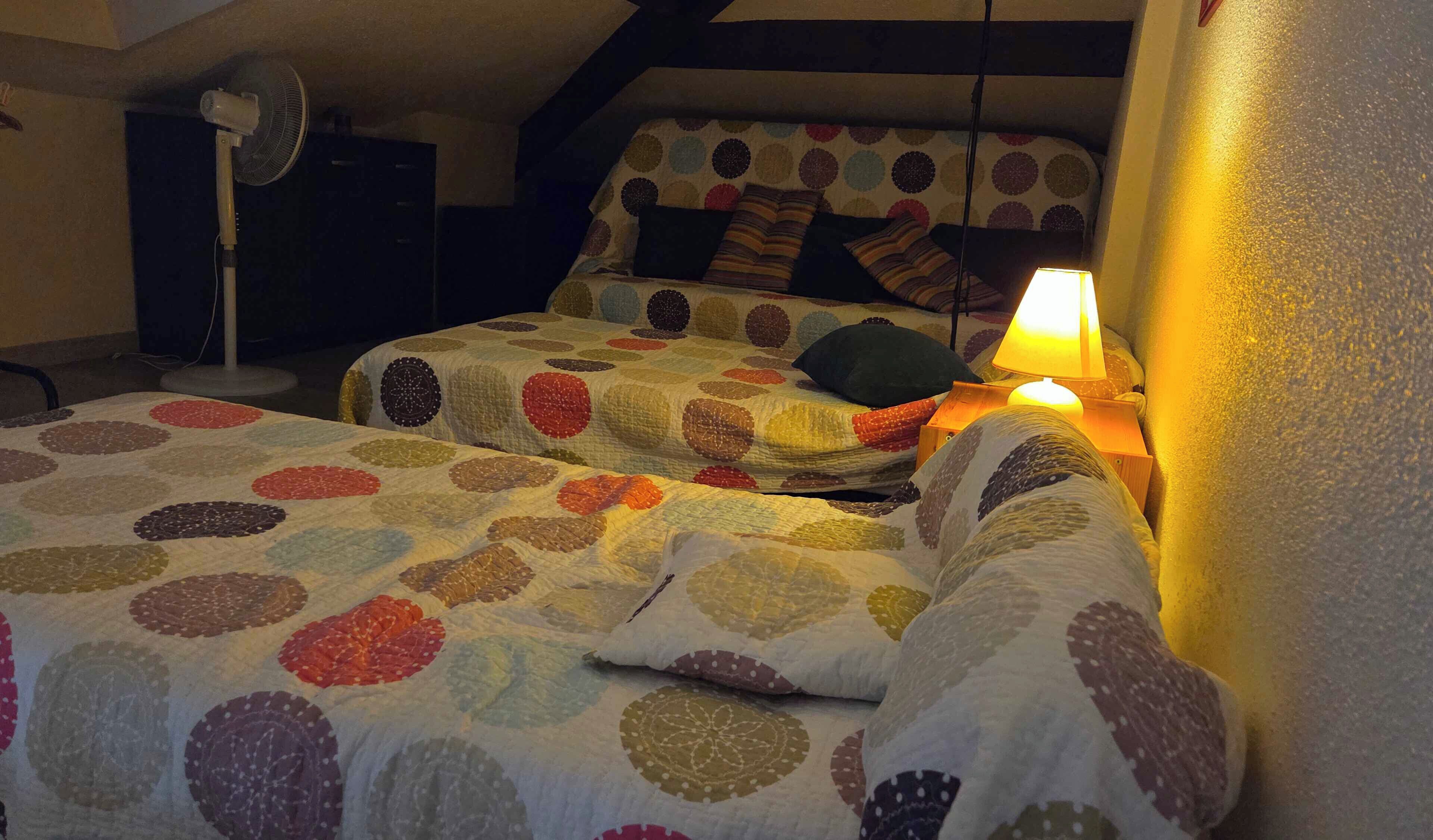 Une pièce chaleureuse avec deux lits recouverts de couvertures aux motifs colorés, un ventilateur sur pied et une lampe diffusant une lumière douce.