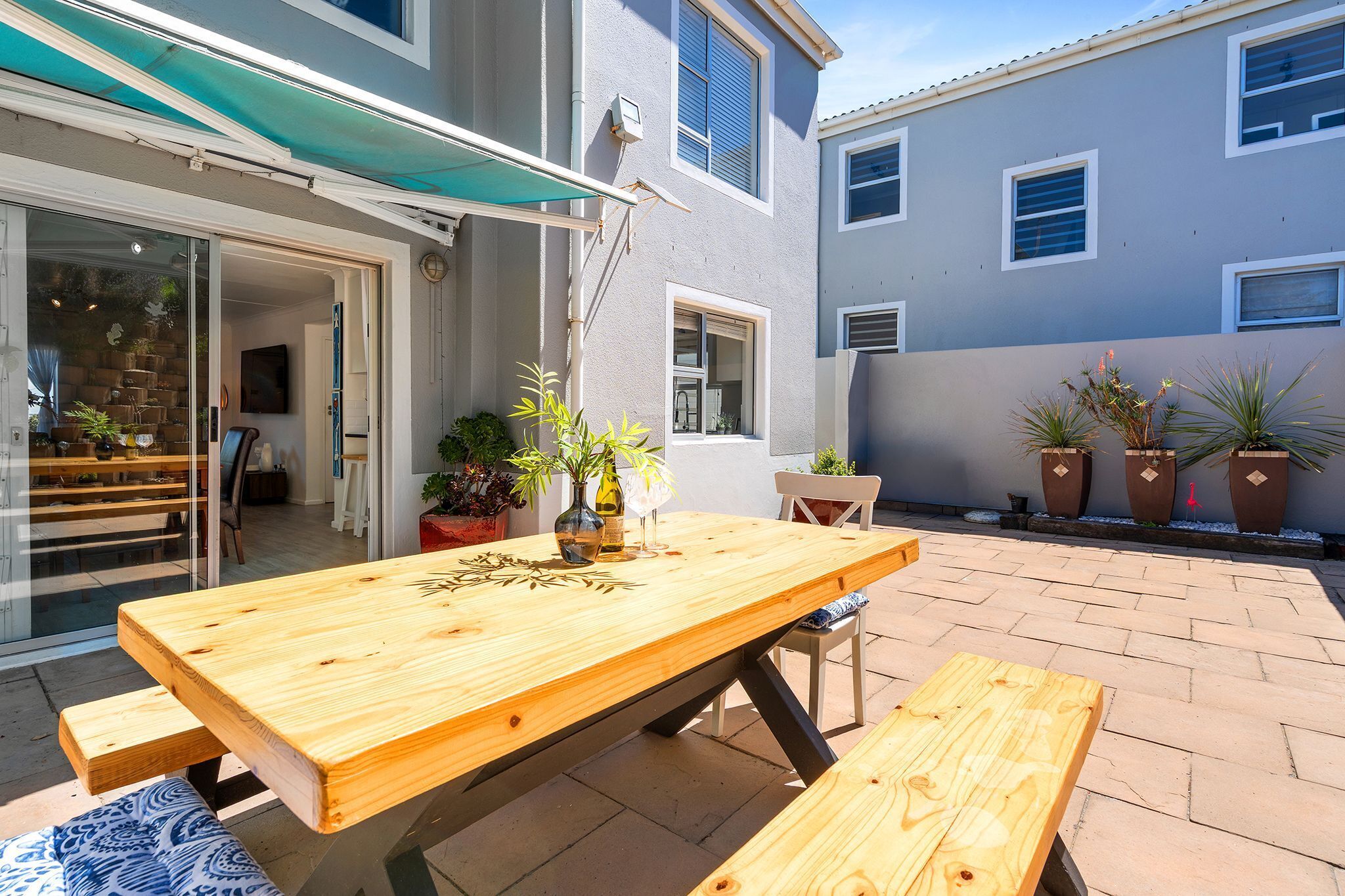 A wooden patio table with benches, potted plants, and a glass door leading to an interior space.