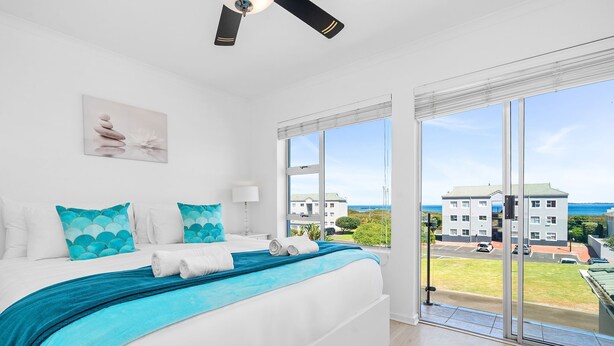 Un dormitorio con cama, ventilador de techo, ventana con vistas a edificios y al mar, y un cuadro en la pared.